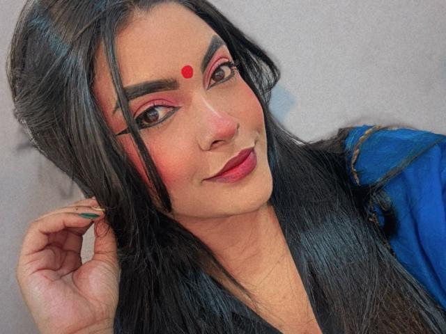 LakshmIrani with dark black hair, pink eye makeup, red dot on forehead, blue dress, and red lipstick, holding her hair, looking slightly to the side.