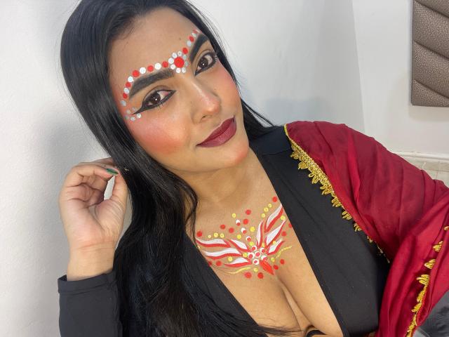 LakshmIrani has dark hair, bold eyebrows, red lipstick, and facial paint with white and red dots. She's wearing a black top, red and gold draped garment, showing her tits.