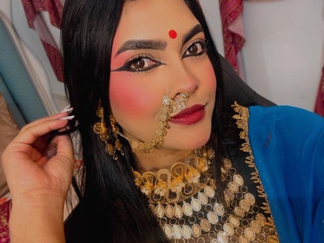 LakshmIrani with black hair, red bindi, gold nose chain, and blue outfit, has pink eyeshadow, red lipstick, and a gold necklace. She's holding her hair, smiling.