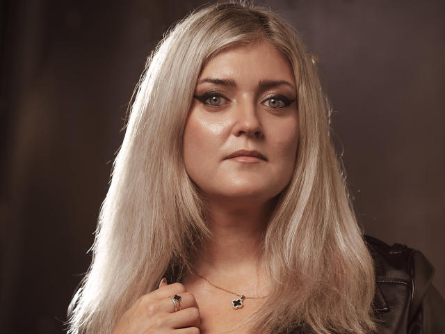 HelenUnique has long, straight blonde hair, blue eyes, and a serious expression. She's wearing a black leather jacket and a small necklace with a black pendant.