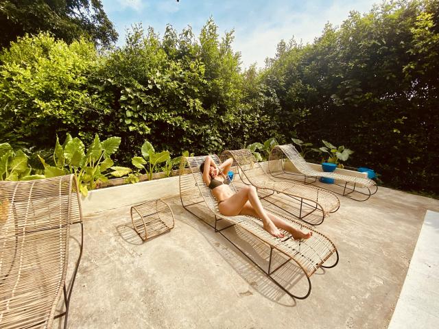 AdharaCooperr liegt auf einer Korbschaukel, trägt einen grünen Bikini, ihr Hintern ist angehoben, die Beine sind gekreuzt, sie hält ein Getränk, das Sonnenlicht wirft Schatten, im Hintergrund ist Grünzeug zu sehen..