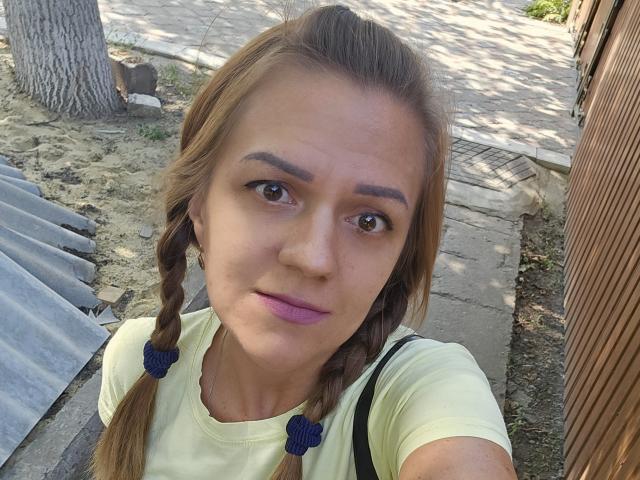 MargaritaNettie with braided brown hair, purple lipstick, and yellow shirt, has brown eyes and a neutral expression, standing outside with a wooden structure on the right.