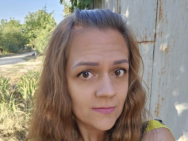MargaritaNettie has wavy brown hair, brown eyes, and pink lipstick, with subtle makeup and a neutral expression, standing near a rusty, white wooden wall.