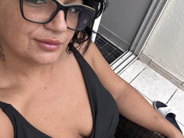YnnaForU with glasses, short black hair, and a black tank top showing her tits. She's sitting, looking at the camera, with a white tile floor and door.