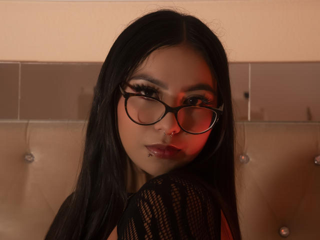 ChloeMoretti with long black hair, black glasses, and red lipstick, looking over her shoulder, showing a pierced nose and dark fishnet sleeve, in a sultry, red-lit photo.