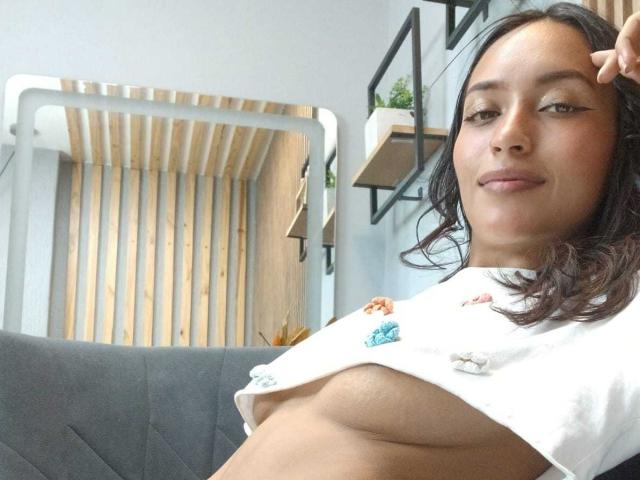 NicolleGlory with medium brown skin, black shoulder-length hair, and a slight smile, lifting her white shirt to expose her small, brown, bare tits. She's sitting on a grey couch.