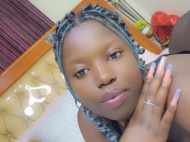 BellaMorgans has dark skin, blue braided hair, and is showing off her glossy pink lips and long, pink-nail-painted fingers with a ring. Her ass is in the background.
