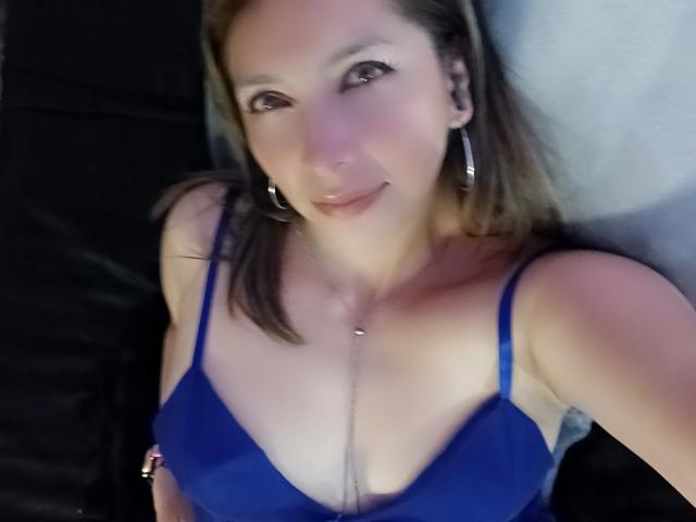 MichelleDumont met een blauwe bh, kleine borsten, lichte huid, bruin haar, oorringen, een ketting, lachend, liggend, zwarte en grijze achtergrond, lichte blos, subtiele make-up, ontspannen..