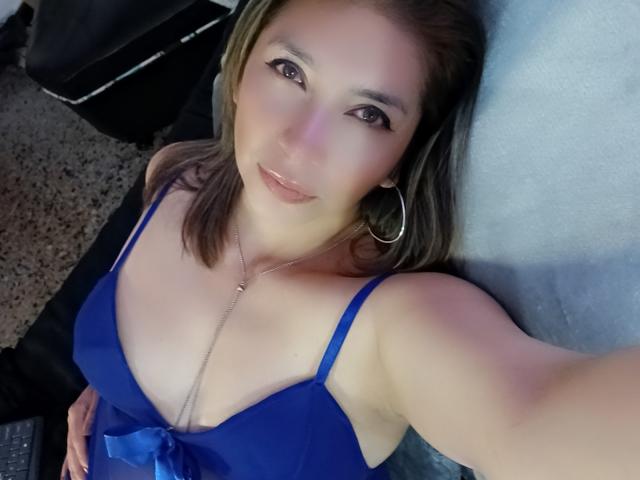 Michelle Dumont, com seios médios, vestindo um vestido azul, deitada, sorrindo, olhos castanhos, brincos de argola, colar, pele clara, cabelo castanho, selfie, laptop no canto inferior esquerdo, carpete ao fundo..