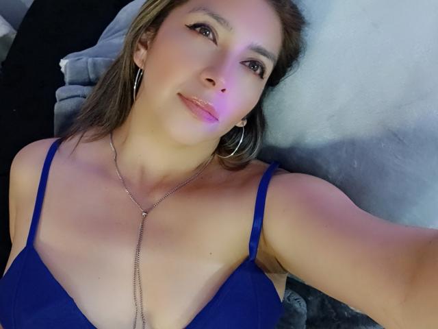Michelle Dumont, com um sutiã azul, seios pequenos, cabelo castanho, brincos de argola, maquiagem discreta e um colar de prata, deitada, sorrindo suavemente, com uma luz rosada-roxa em seu rosto..