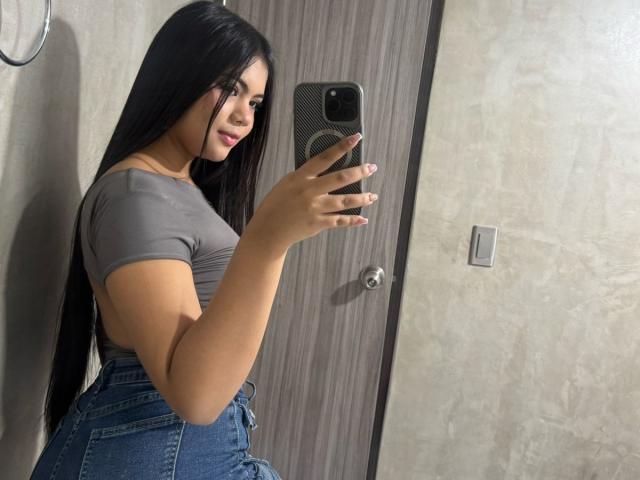 AbbyColinss with long black hair, gray tight shirt, and blue jeans, holding a phone, showing her curvy ass and small tits in a mirror selfie.