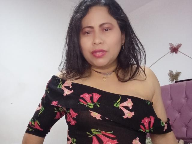 MiaSalomeChaude with medium brown skin, black hair, pink floral off-shoulder black dress, pink lipstick, necklace, and purple headboard in background. Boobs visible through dress.