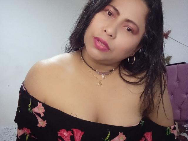 MiaSalomeChaude con pechos medianos, vestido floral negro, lápiz labial rosa, aros grandes, collar y cabello negro largo, inclinada hacia adelante, sonriendo ligeramente..