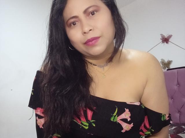 MiaSalomeChaude, con cabello negro largo, lápiz labial rosa y una blusa negra con flores y corte que deja los hombros al descubierto, mostrando sus senos, lleva un collar y sonríe ligeramente en un dormitorio..