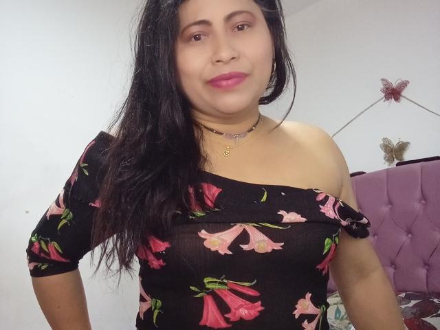 MiaSalomeChaude, com cabelos longos e pretos, batom rosa, vestido floral preto com um ombro descoberto, um colar de ouro pequeno, sorrindo, pele marrom clara, seios de tamanho médio, em pé em um quarto..