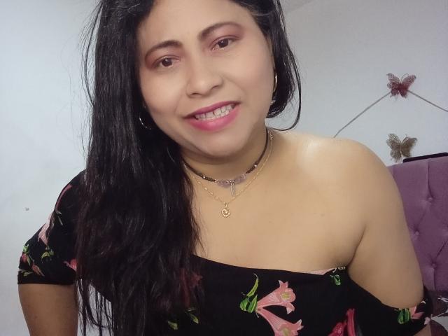 MiaSalomeChaude, com cabelos longos e pretos, batom rosa, vestido floral preto com um ombro descoberto, colar de ouro, sorrindo, mostrando os seios, mamilos rosa não visíveis, em um quarto..