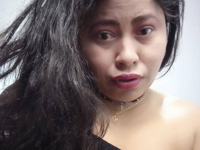 MiaSalomeChaude a les cheveux noirs et ondulés, du rouge à lèvres rose et une expression légèrement confuse. Elle porte un bandeau noir et un collier en or avec un petit pendentif..