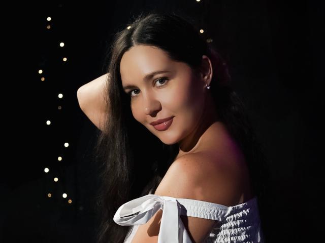 LucyBi, con lunghi capelli neri, una maglietta bianca a spalle scoperte, piccoli orecchini, sorridente, con un braccio alzato, sfondo scuro con piccole luci bianche, luce soffusa sul viso. Il seno è parzialmente visibile..