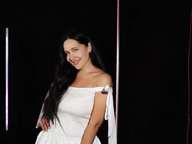 LucyBi met lang zwart haar, lachend, gekleed in een witte jurk met een open schouder, staat voor een donkere achtergrond met roze en witte neonlichten aan de linkerkant. Borsten zichtbaar..
