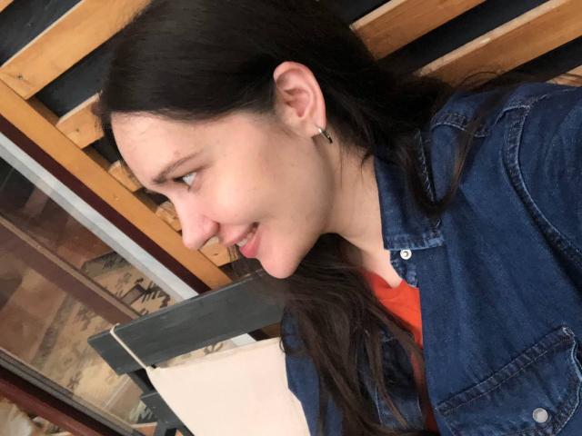 LucyBi met lang donker haar, een lichte glimlach, een blauwe denim jas, kleine oorbellen, lichte huid, profiel van de zijkant, kijkt naar links, draagt een rood shirt, een kleine puist op de wang..