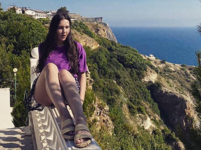 LucyBi with long brown hair, purple shirt, black skirt, and sandals, sitting with legs crossed, showing her ass, on a white railing, overlooking a blue ocean.