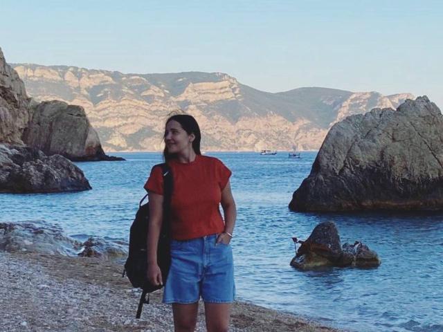 LucyBi with brown hair, red shirt, and blue shorts stands on rocky shore, hand in pocket, black backpack, looking at water with rocks and mountains in background.