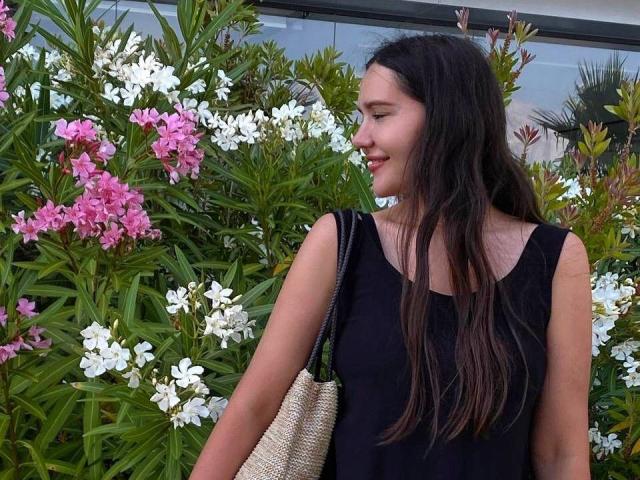 LucyBi mit langen, braunen Haaren, einem schwarzen Top ohne Ärmel, lächelnd, hält eine beige geflochtene Tasche und steht vor rosa und weißen Blumen. Die Brüste sind nicht sichtbar..