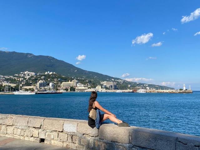 LucyBi est assise sur un mur de pierre, le dos à la caméra, vêtue d'une robe bleue, avec des cheveux bruns, un petit sac en cuir marron, les jambes croisées, et elle regarde la mer bleue et la ville lointaine..