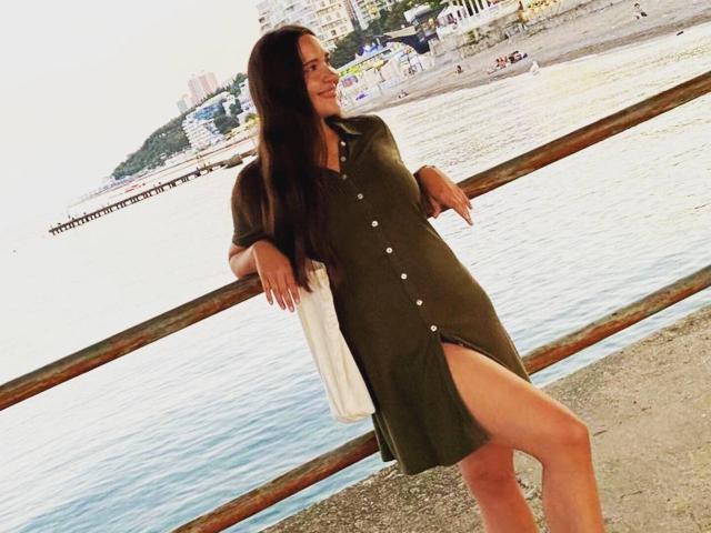 LucyBi, avec ses longs cheveux noirs, sa robe verte avec des boutons blancs et une fente sur la cuisse, est appuyée contre une rambarde en bois près de l'eau, et elle sourit. Ses fesses sont légèrement visibles de côté..