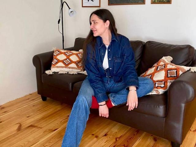 LucyBi, con cabello castaño, una chaqueta vaquera y jeans azules, está sentada en un sofá de cuero marrón, con las piernas separadas, sonriendo y mostrando su trasero con unos pantalones rojos, sobre un piso de madera..