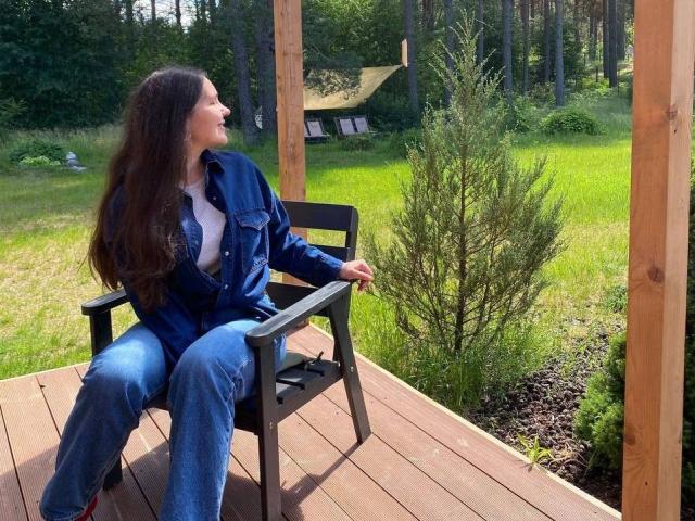 LucyBi, avec ses longs cheveux bruns, est assise sur une chaise en bois, vêtue d'une chemise en jean bleu et d'un jean, et regarde vers la droite, sur une terrasse en bois baignée de soleil..