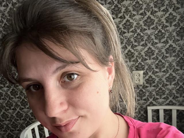 ToniElizabeth has light brown eyes, a small nose, and a slight smile. She's wearing a pink shirt, has a small beauty mark on her cheek, and a single earring.