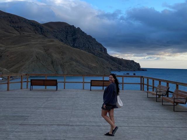 LucyBi se tient sur une promenade en bois, avec l'océan bleu et des collines rocheuses en arrière-plan. Elle porte une chemise sombre, un short noir et des chaussures noires, et tient un sac blanc..