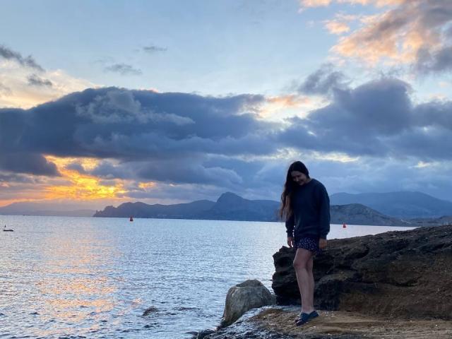 LucyBi, avec ses longs cheveux bruns, se tient sur une plage rocheuse, vêtue d'un sweat à capuche sombre et d'un short à motifs léopard, le regard baissé, avec un ciel de coucher de soleil aux nuages orange et bleus en arrière-plan..