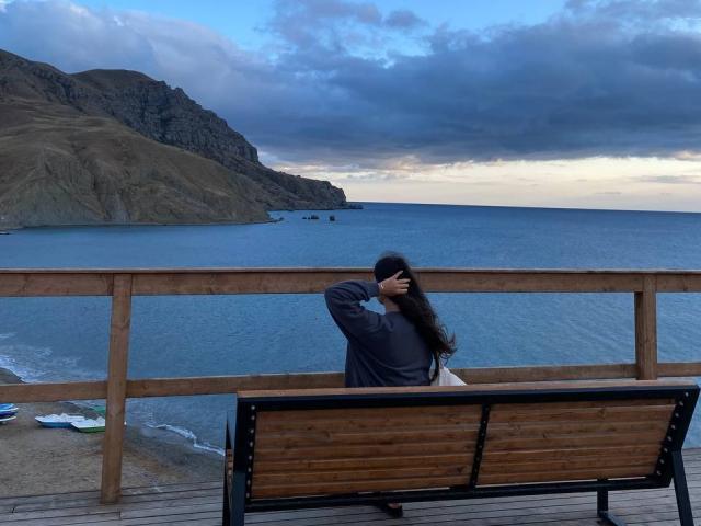 LucyBi, com cabelos longos e escuros, vestindo um moletom cinza, está sentada em um banco de madeira, com a mão no cabelo, de frente para o oceano azul, céu nublado, uma colina rochosa à esquerda e um corrimão de madeira..