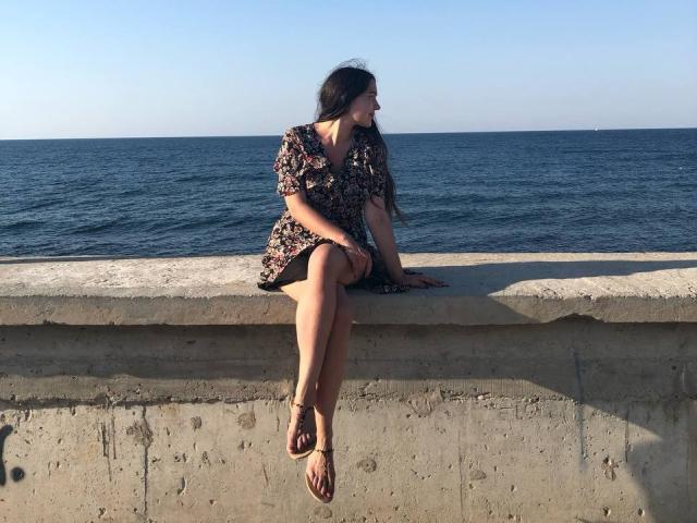 LucyBi, avec ses longs cheveux foncés, sa robe fleurie et ses sandales, assise sur un mur en béton, les jambes croisées, regardant l'océan, un ciel bleu, en journée, décontractée, détendue, petite poitrine, mignonne..