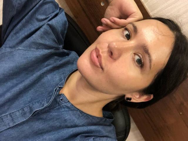 LucyBi, con un seno di media grandezza, indossa una camicia blu con bottoni, ha dei cerchi all'orecchio neri, occhi marroni, labbra rosa leggermente dischiuse, capelli scuri, una mano appoggiata sulla testa, è seduta indietro su una sedia nera..