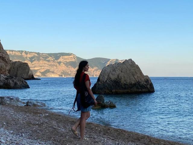 LucyBi, com cabelos longos e pretos, camisa vermelha, shorts de jeans e uma bolsa preta, caminhando descalça em uma praia rochosa, olhando para o oceano azul com grandes rochas e montanhas distantes..