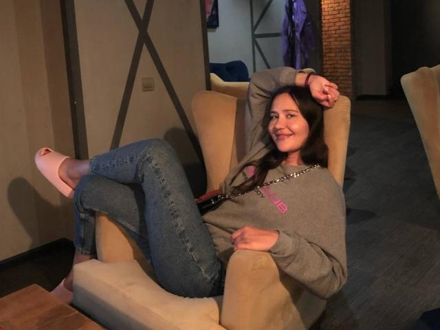 LucyBi est assise dans un fauteuil beige, vêtue d'un pull gris, d'un jean bleu et de talons roses, avec son bras derrière la tête et un sourire détendu..