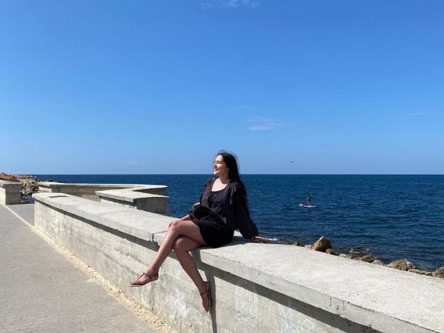 LucyBi, con cabello y vestido negros, está sentada en un muro de concreto, con las piernas cruzadas, con el océano azul y el cielo despejado al fondo, usando sandalias marrones, mirando hacia la derecha..
