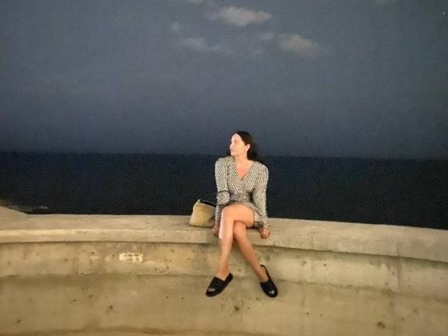 LucyBi, con un vestito a pois, scarpe nere e una borsa color cuoio, è seduta sul cemento, con le gambe incrociate, e guarda verso il cielo serale di un blu scuro. Il suo sedere è leggermente visibile, e ha i capelli scuri..