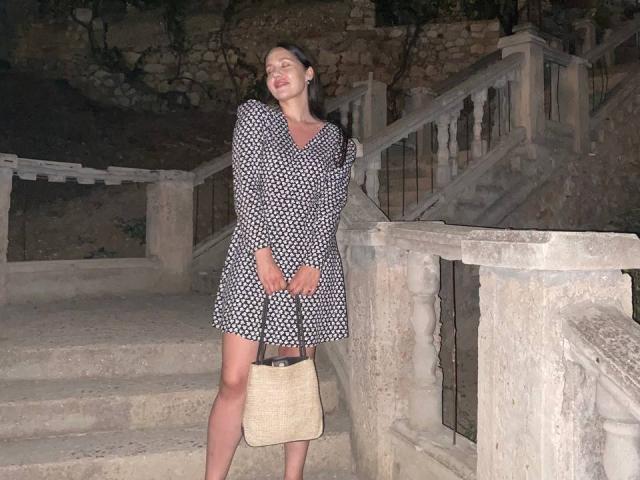 LucyBi è in piedi su delle scale di pietra, indossa un vestito a pois bianchi e neri, tiene una borsa beige, ha gli occhi chiusi e un leggero sorriso. Ha il seno..