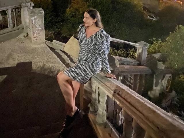 LucyBi in a black and white polka dot dress, sitting on a wooden railing at night, legs crossed, black boots, brunette, smiling, hand on railing, looking down.