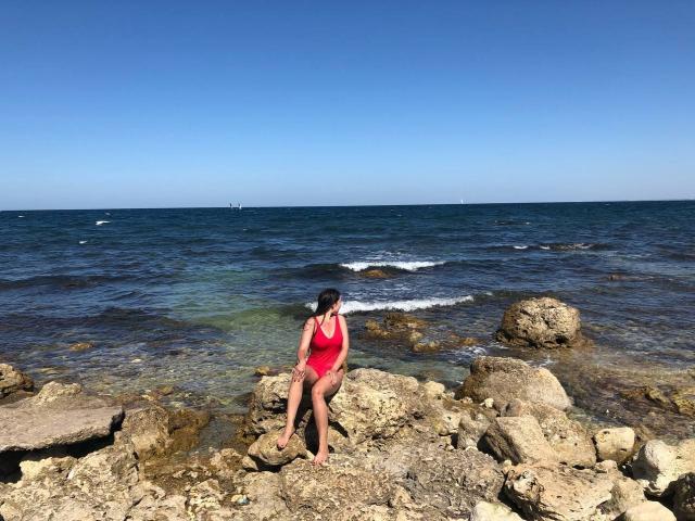 LucyBi mit schwarzen Haaren, in einem roten Badeanzug, sitzt auf einer felsigen Küste, die Beine verschränkt, im Hintergrund das Meer und ein klarblauer Himmel, in der Ferne ein kleines Boot auf dem Wasser..
