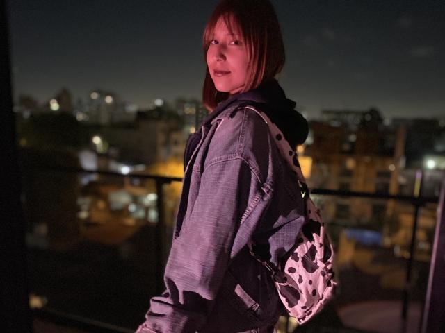KiaraHernandezX with red-brown hair, wearing a gray hoodie and black-and-white backpack, standing in a dimly lit urban night setting, looking back with a slight smile.