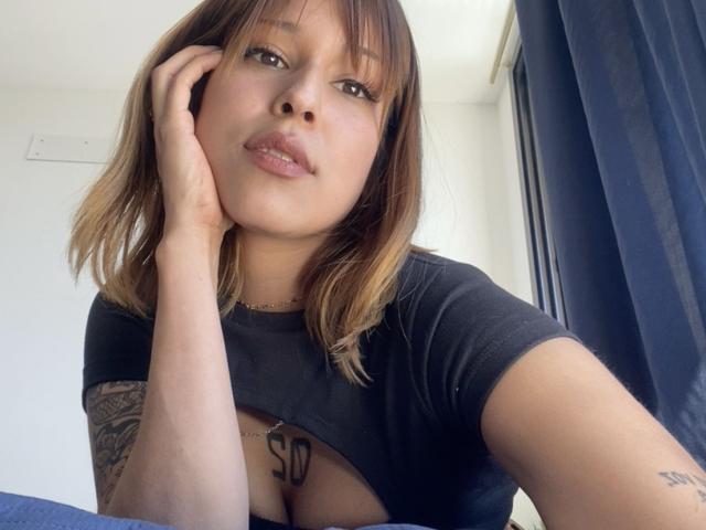 KiaraHernandezX with shoulder-length brown hair, black top, and tattoo on chest, resting chin on hand, looking at camera, showing small tattoos, and subtle cleavage.