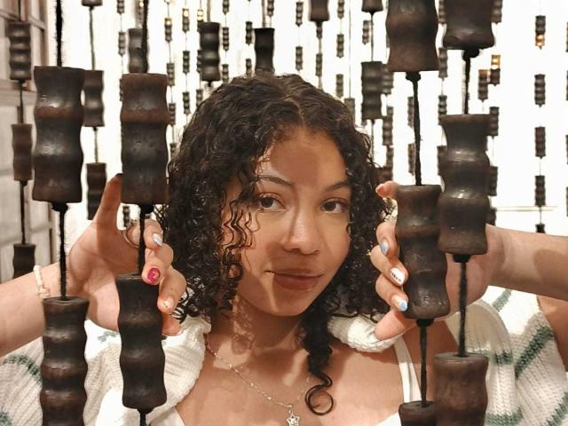 GaiaBlar, con i capelli neri ricci, tiene dei bastoncini di legno marrone scuro davanti al viso, indossa una maglietta bianca e una collana, e sorride dolcemente..
