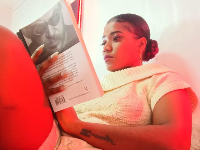 Caylin with dark skin, tied-up hair, and white knit sweater, reading a book, showing her tattooed arm and red-lit butt, holding the book near her face.