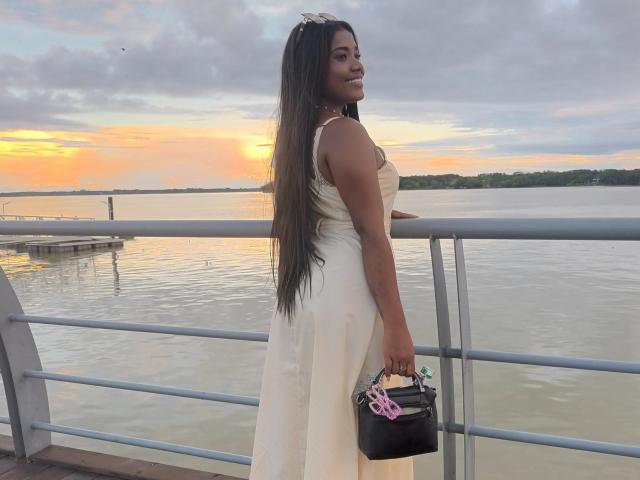 Caylin with long brown hair, tan skin, wearing a white dress, holding a black purse with pink straps, standing on a waterfront railing at sunset.