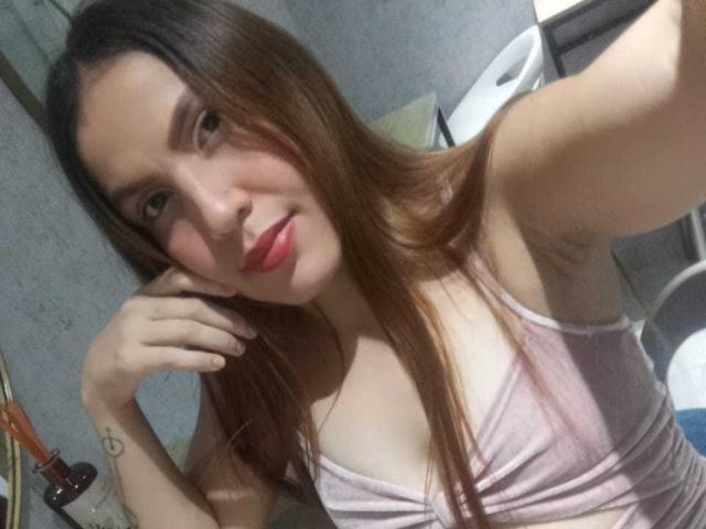 SusanThomass with long brown hair, pink lipstick, and a pink top, showing her tits and a small tattoo on her right arm, taking a selfie with a relaxed smile.